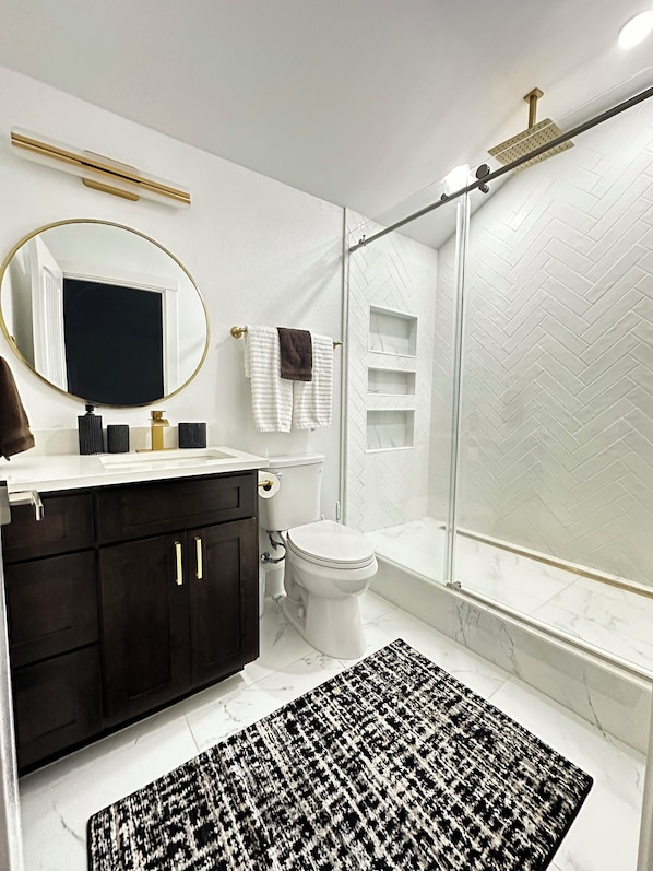 A modern bathroom with a white herringbone tile backsplash, a black and white patterned rug, a gold-framed mirror, and a glass-enclosed shower. - Redlands farm (Oregon City)