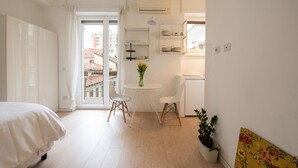 Een lichte, compacte kamer met een bed, een eettafel met stoelen, een plant in pot en uitzicht op gebouwen.