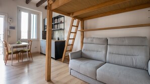 Una habitación con techo de madera, un sofá gris, una mesa de comedor con sillas y una zona de cocina con estantes y una escalera.