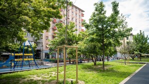 Leikkikentällä on keinut ja kiipeilyteline, ja se on ympäröity ruoholla ja puilla. Taustalla näkyy rakennus.