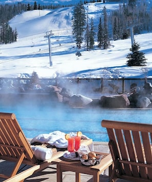 A wooden deck with a table set for breakfast, overlooking a hot spring and a snowy mountain landscape.