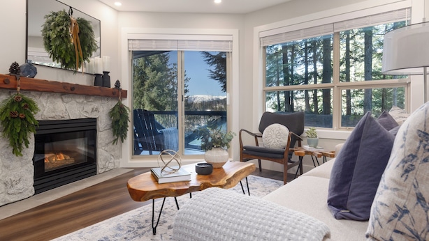 A cozy living room with a fireplace, a large window with a forest view, a comfortable sofa, and a wooden coffee table.