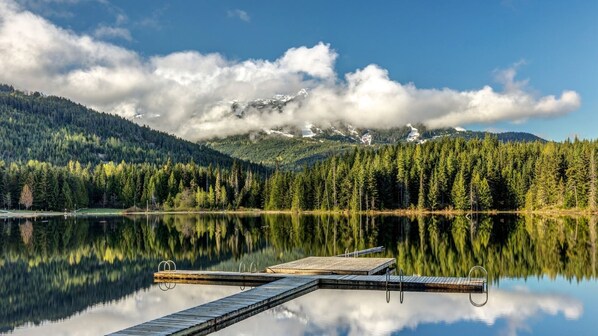 Un lago sereno con un muelle de madera, rodeado de un bosque denso y montañas nevadas al fondo.
