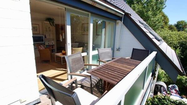 Un patio al aire libre con una mesa y sillas de madera, una puerta corrediza de vidrio que da a un espacio interior con un televisor y estanterías, y un techo inclinado.
