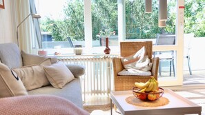 Una sala de estar moderna con un sofá, una silla de mimbre, una mesa con frutero y ventanas amplias con vista a zonas verdes.