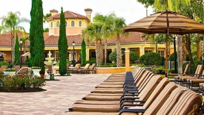 Un espace aménagé au bord de la piscine, avec des chaises longues et des parasols, une fontaine et un bâtiment doté d’un toit recouvert de tuiles.
