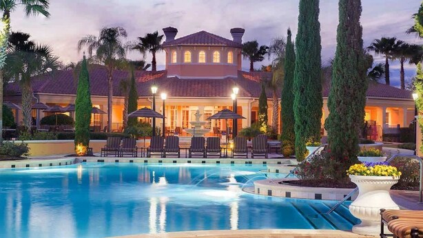 Une piscine avec des chaises longues et des parasols, une grande maison avec une fontaine et de grands arbres.