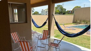 Uma varanda coberta com quatro cadeiras de descanso listradas, uma rede e vista para uma piscina e vegetação.