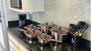 Uma cozinha com bancada preta, panelas de aço inoxidável com tampas, cafeteira e porta-utensílios.