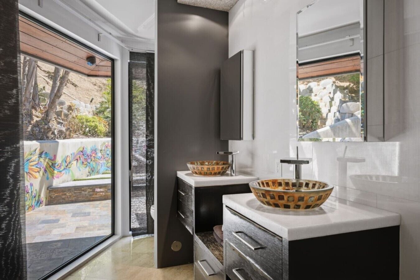 A modern bathroom with a large window, two sinks with decorative bowls, and a dark cabinet.