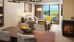 Une chambre d’hôtel avec une cheminée, un coin repas avec une table et des chaises en bois, un canapé, une télévision et un balcon offrant une vue sur les montagnes.