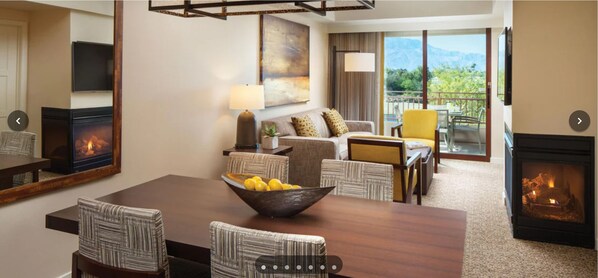 Habitación de hotel con chimenea, zona de comedor con mesa y sillas de madera, sofá, televisor y balcón con vistas a las montañas.