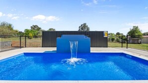 A swimming pool with a waterfall feature, surrounded by a fence and a wooden structure in the background. - Bellevue Holiday House (Moore Creek)