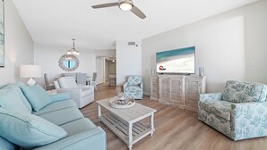 A spacious living room with a ceiling fan, a television mounted on the wall, and a large window. - BEACHFRONT, FAMILY FRIENDLY, INDOOR/OUTDOOR POOL & HOT TUB (Orange Beach)