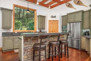 A kitchen with a stone island, bar stools, a stainless steel refrigerator, and a large window overlooking a forest.
