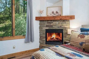 A room with a stone fireplace, a bed with patterned bedding, a window with a view of greenery, and a framed picture above the fireplace.