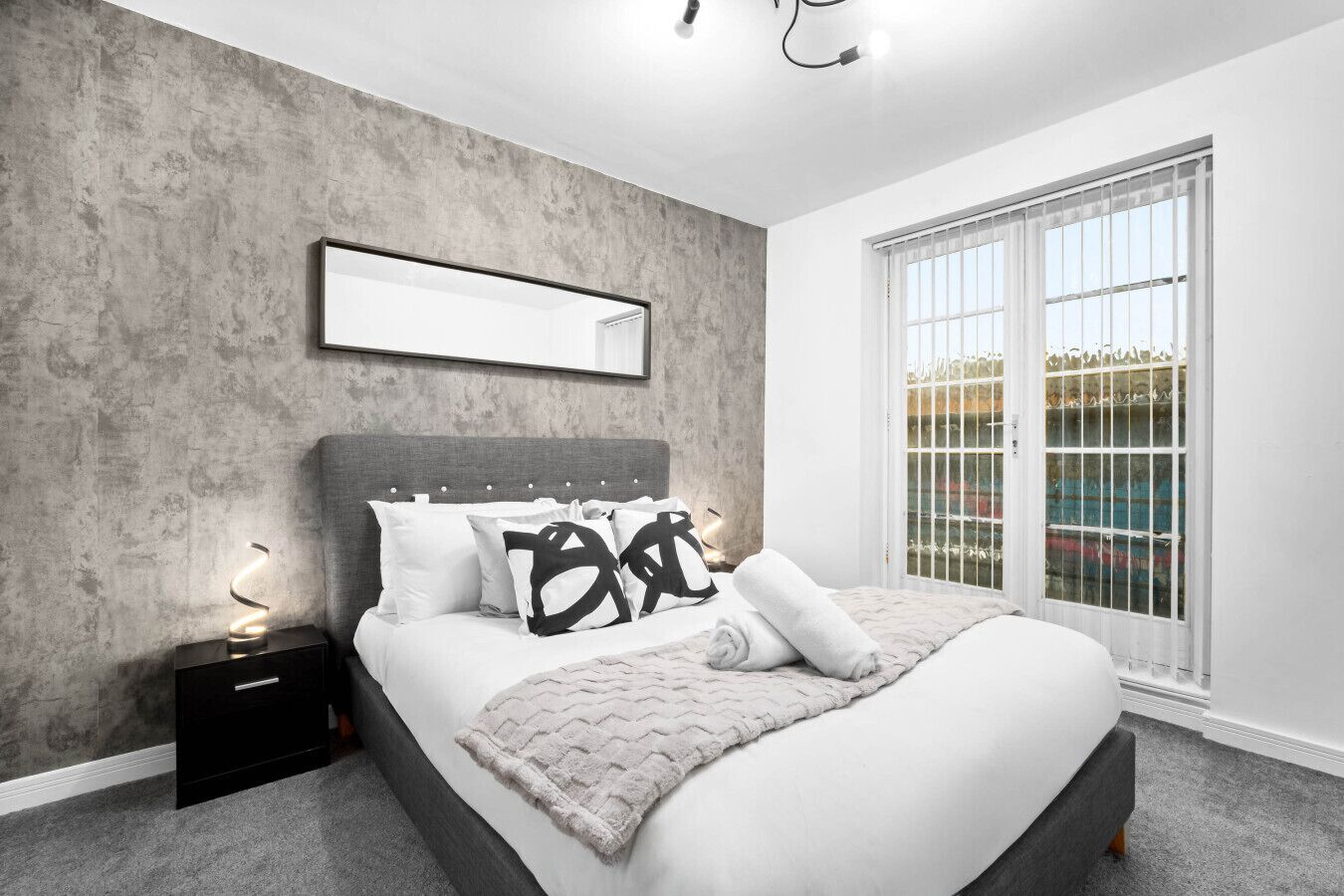 A modern bedroom with a large bed, grey concrete walls, a window with a view, and a bedside table with a lamp.