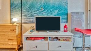 A room with a large wave painting, a TV on a white cabinet, a red chair, and a small table with a lamp. - Primavéra by Interhome (Lacanau)