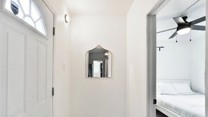 Un pasillo estrecho con una puerta blanca, un espejo pequeño y un ventilador de techo en un dormitorio.