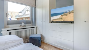 A bedroom with a large window, a bed, a bedside table with a candle, a TV on a stand, and a view of a beach.