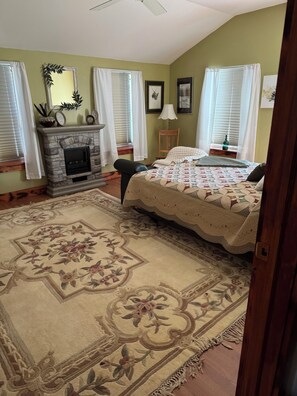 A bedroom with a fireplace, a bed with a floral bedspread, a large rug, and a window with blinds. - Charming 4-bedroom house is located in a quiet, small town of Rossie. (Hammond)