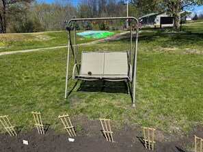 Een metalen schommelstoel met een netzitplaats en -rugleuning, geplaatst op een gazon met een onverharde pad in de buurt.