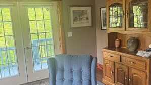 Una habitación con un sillón azul, vigas de madera en el techo, un ventilador de techo, un aparador de madera con puertas de cristal y un cuadro en la pared.