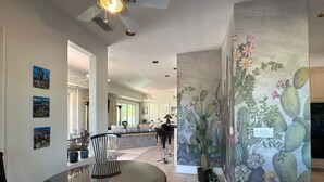 A spacious living area with a round glass table, a ceiling fan, and a cactus-themed mural.