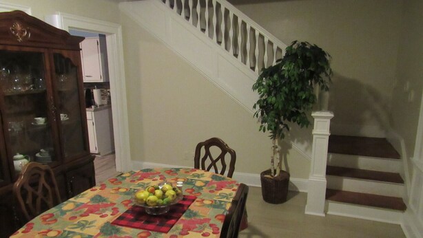 A dining area with a table covered by a fruit-patterned tablecloth, a wooden chair, a potted plant, and a staircase in the background.