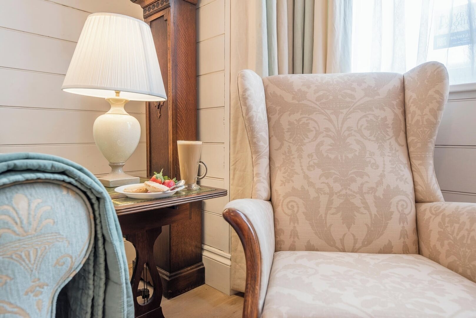 Una habitación con un sillón estampado, una mesita de madera con una lámpara, un plato de comida y un vaso de bebida.