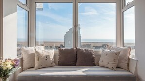 Une pièce dotée d’une grande fenêtre donnant sur la plage et un bâtiment. On y trouve un banc rembourré avec des coussins et un vase rempli de fleurs.