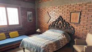 Quarto com parede de tijolos, cama com colcha estampada, uma cadeira, um banco com almofadas amarelas e uma janela com persianas.