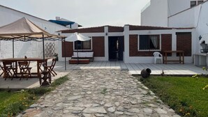 Um pátio com caminho de pedra, mesa e cadeiras de madeira, uma tenda branca e um edifício com fachada de tijolos e grandes janelas.