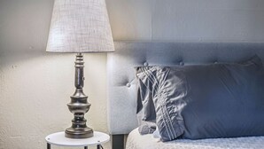 A bed with a grey headboard and pillows, a bedside table with a lamp, and a white wall. - Benedictine Theme 2br 1ba (Atchison)