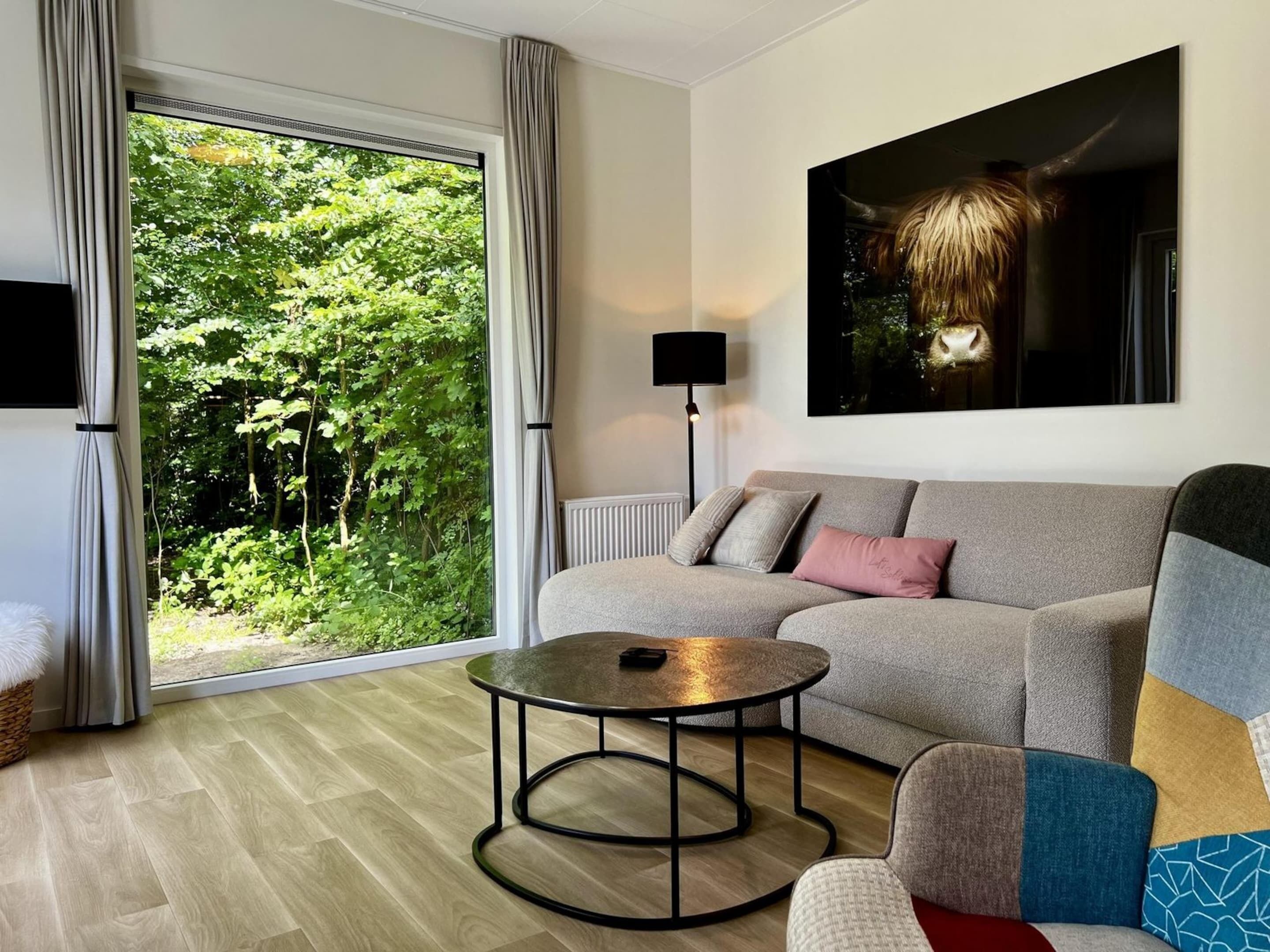 Un salon moderne avec un canapé gris, une table basse ronde et une grande fenêtre donnant sur un paysage verdoyant.