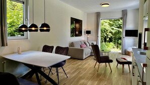 Un salon moderne comprenant un coin repas, un canapé, un téléviseur et une grande fenêtre donnant sur un paysage verdoyant.