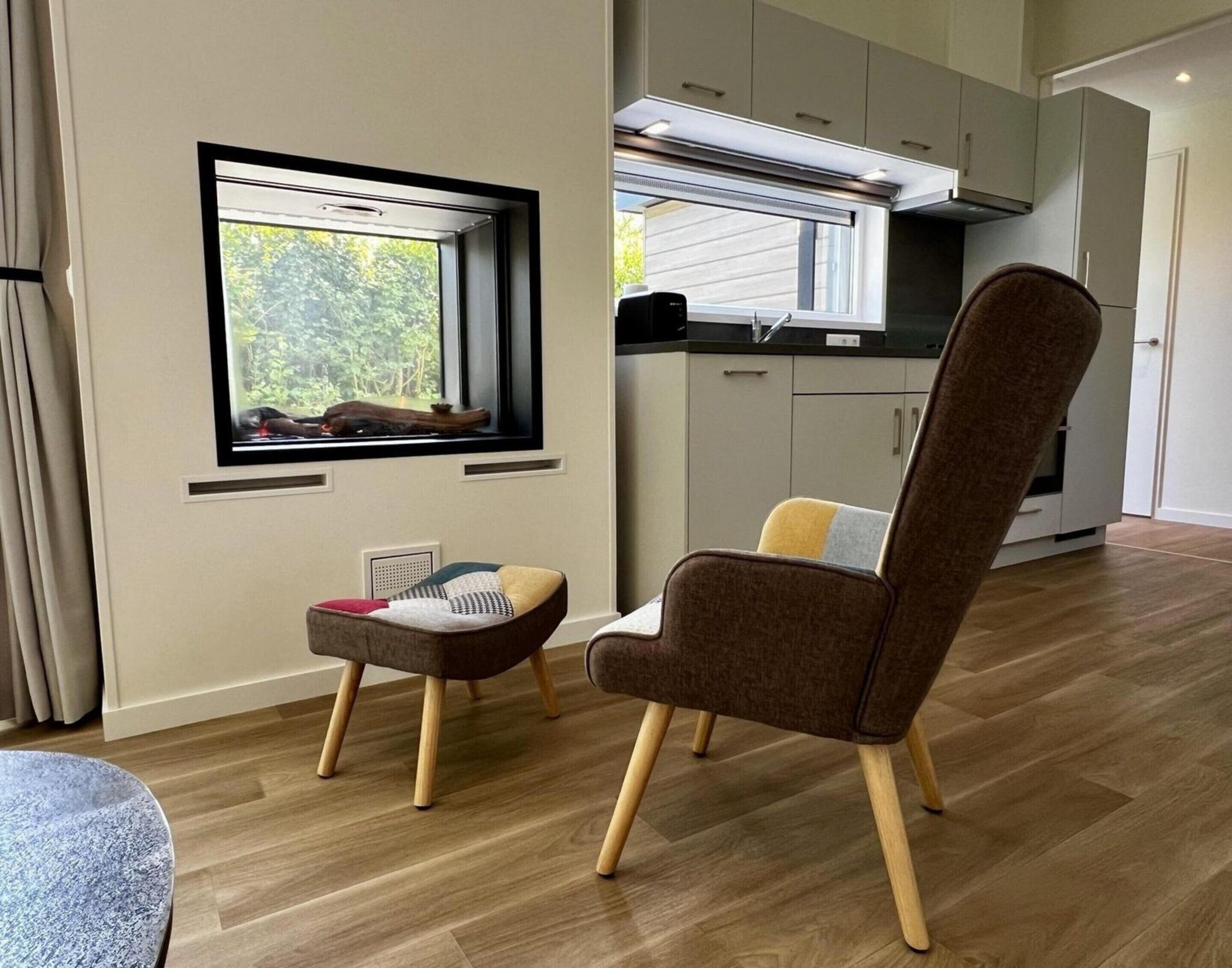 A modern kitchen with white cabinets, a window with a view of greenery, and a brown chair with wooden legs.