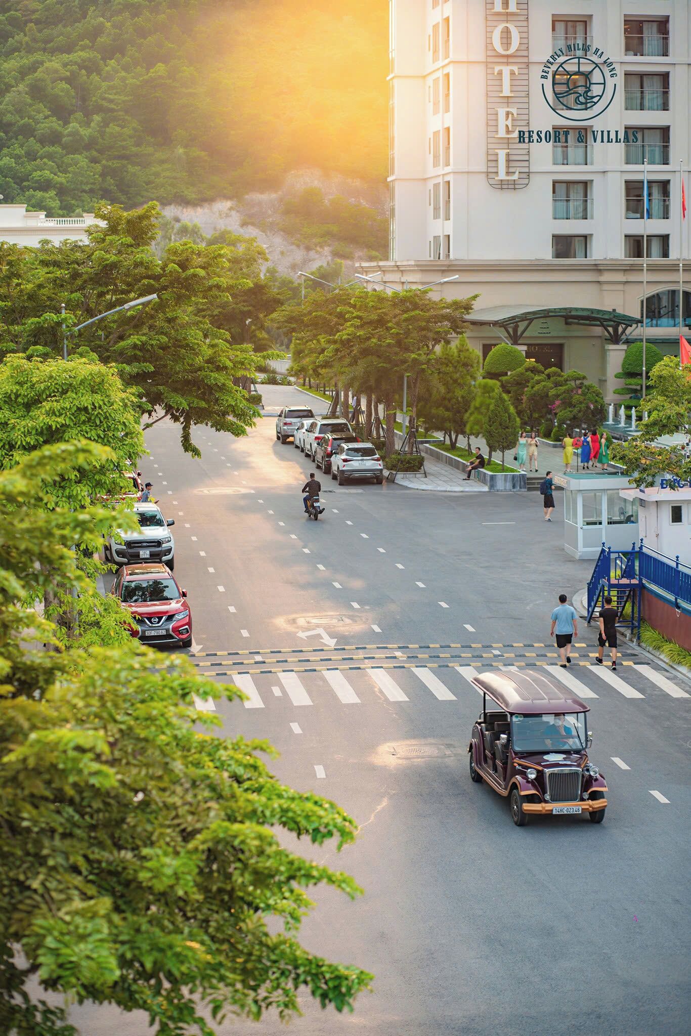 Un hotel con un letrero llamativo, una calle con coches estacionados y un tuk-tuk en la vía.
