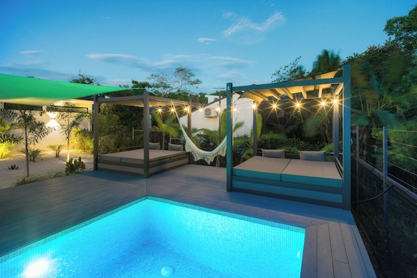 A swimming pool with a wooden deck, a hammock, and a covered seating area with string lights.