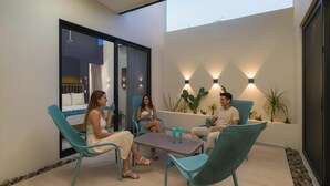 Un área de estar al aire libre moderna con sillas azul verdosas y una mesa, rodeada de plantas en macetas e iluminación instalada en la pared.