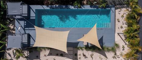 A rooftop pool with clear blue water, surrounded by a sandy area with plants and a shaded structure.