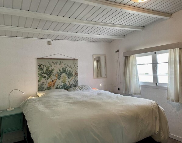 Un dormitorio con cama, mesita de noche, espejo en la pared y ventana con cortinas.