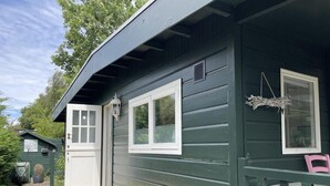 A green wooden building with two windows, a door, and a small porch with a bench. - Furnished Chalet in Sint Maartenszee (Sint Maartensvlotbrug)