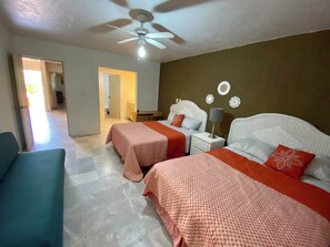 A hotel room with two beds, a ceiling fan, a nightstand with a lamp, and a view of a hallway and another room.
