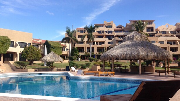 Un área de piscina con sillas de descanso y sombrillas de paja, rodeada de edificios de varios pisos con balcones.
