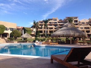 A pool area with lounge chairs and thatched umbrellas, surrounded by multi-story buildings with balconies.