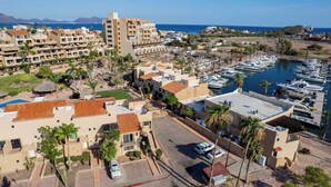 Vista aérea de un pueblo costero con un puerto deportivo, edificios residenciales y un hotel destacado con varios pisos y balcones.