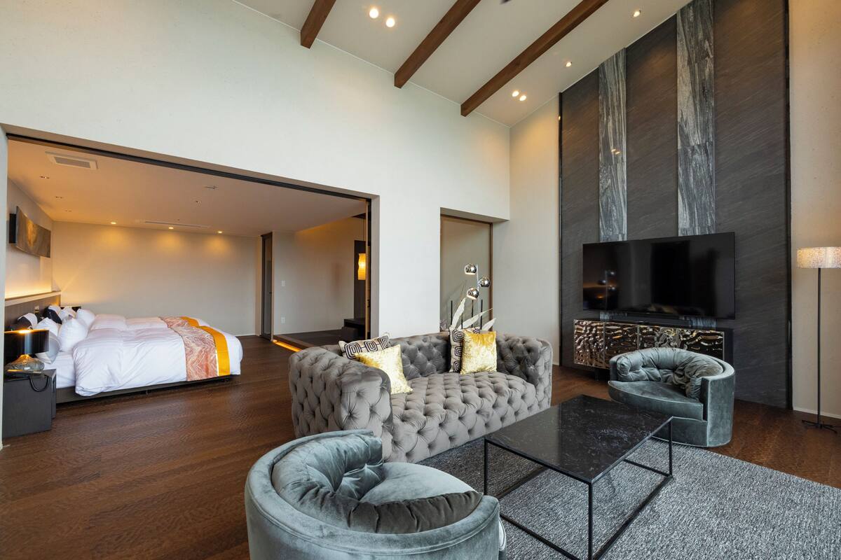 Una habitación de hotel moderna con una cama grande, un sofá gris capitonado, un televisor de pantalla plana y una chimenea.