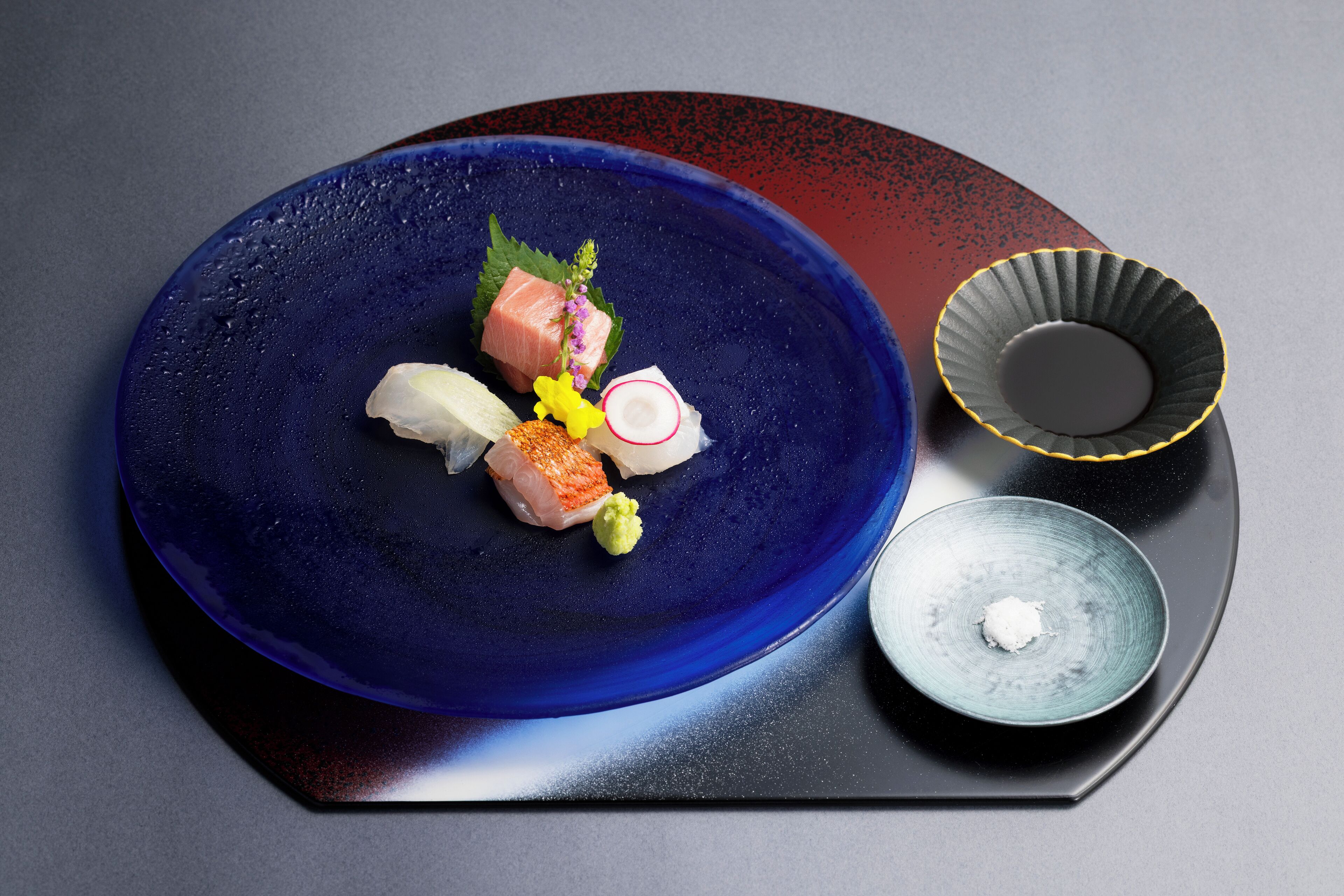 A blue plate with various sushi pieces and a small bowl of sauce on a dark tray.