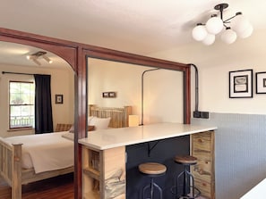 A room with a bed, a desk with stools, a window with curtains, and a ceiling light fixture.
